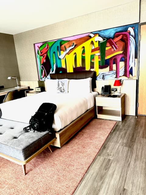 This image depicts a hotel suite complete with a white bed in the center of the room. Atop it are pillows, while a colorful piece of abstract art hangs on the wall above it. There is a wooden side table next to it with a lamp and telephone on top of it. At the end of the bed is a grey bench with a black backpack resting atop it. The bed sits on top of an orange rug and wood paneled flooring.