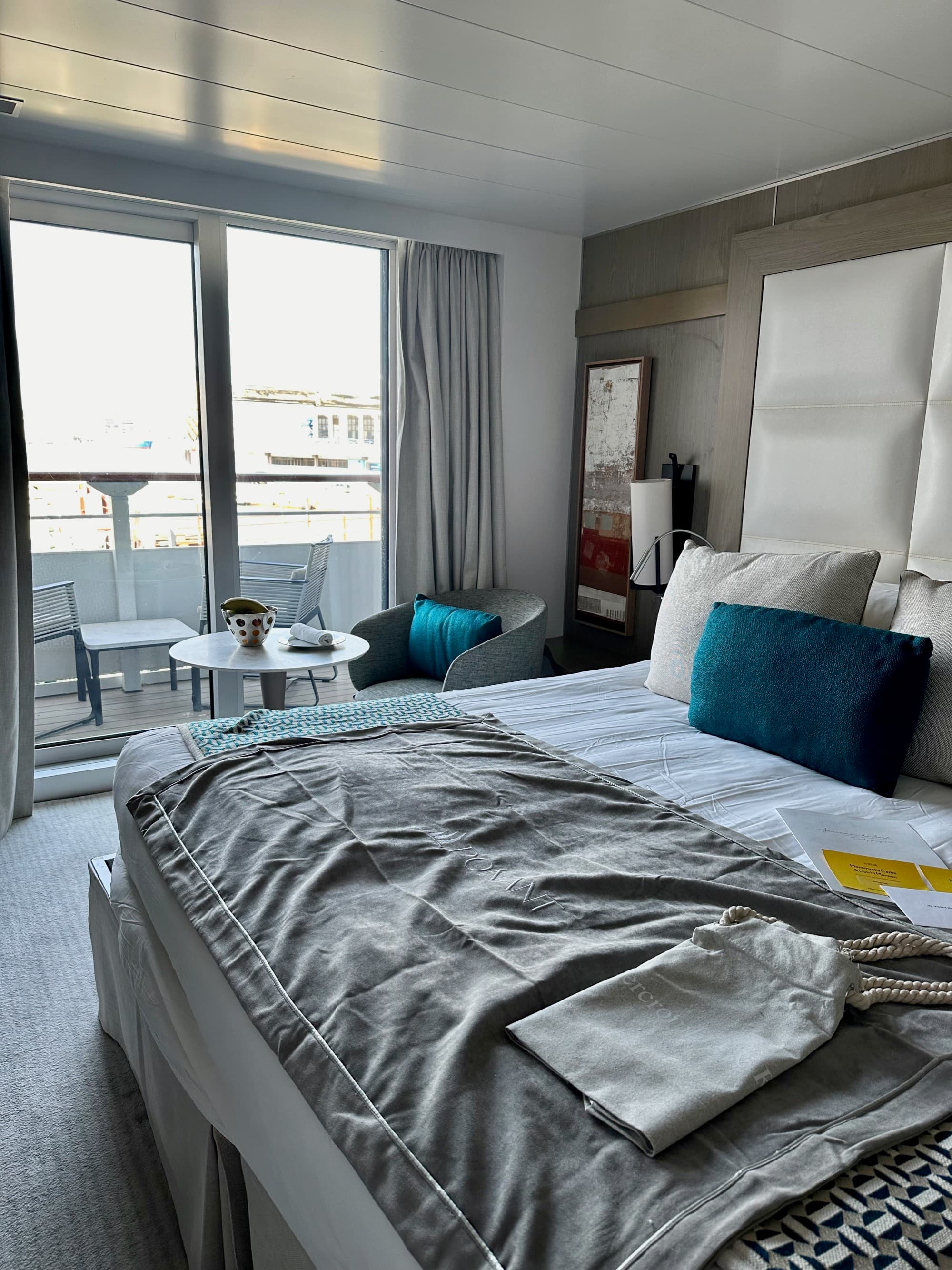 A cabin on the ship with a white bed, gray blanket, blue pillow and balcony - Kristyne Wada