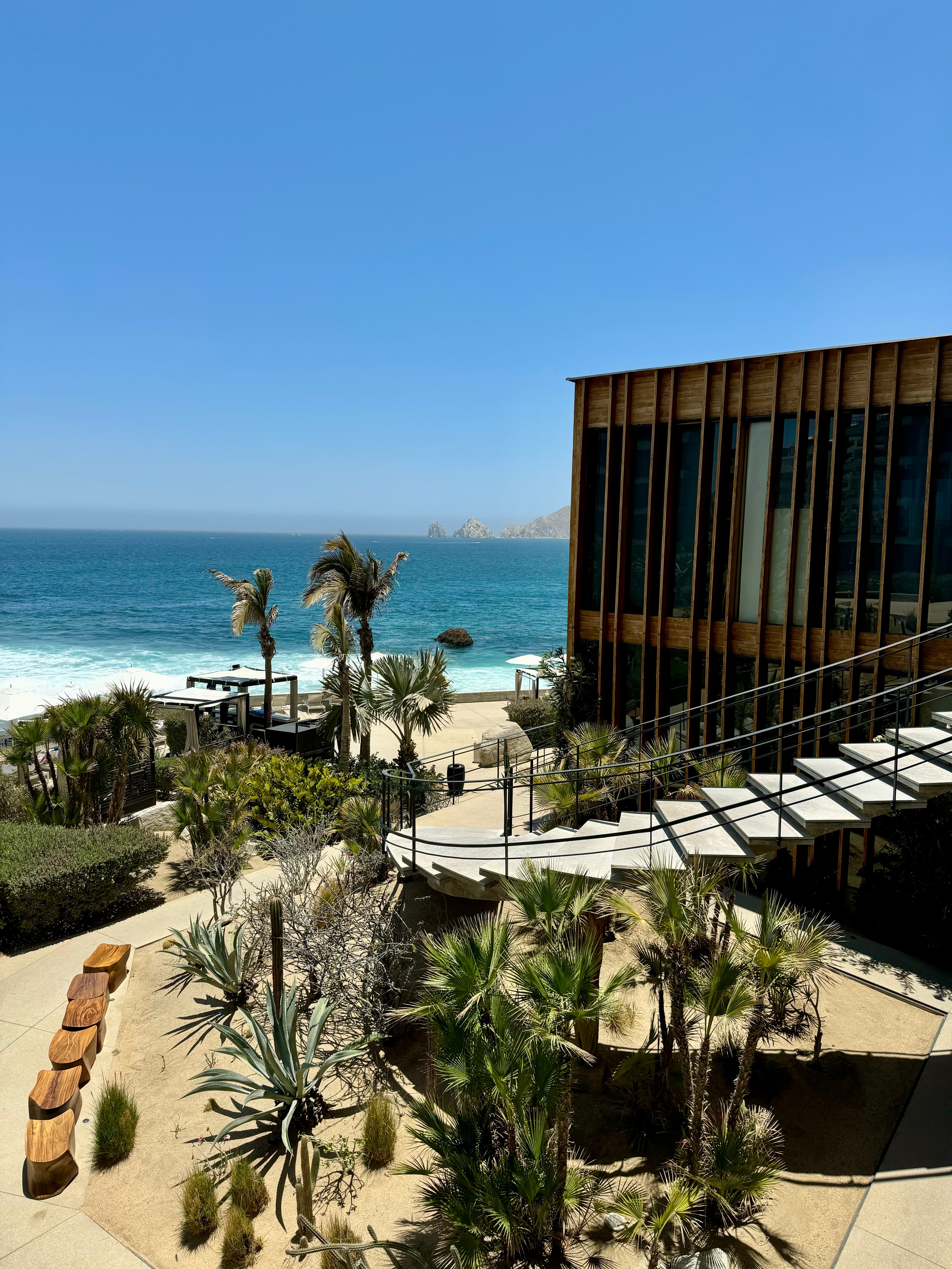 A view of a oceanfront resort with curved stairs leading down to the beach landscaped with palm trees on a sunny day.