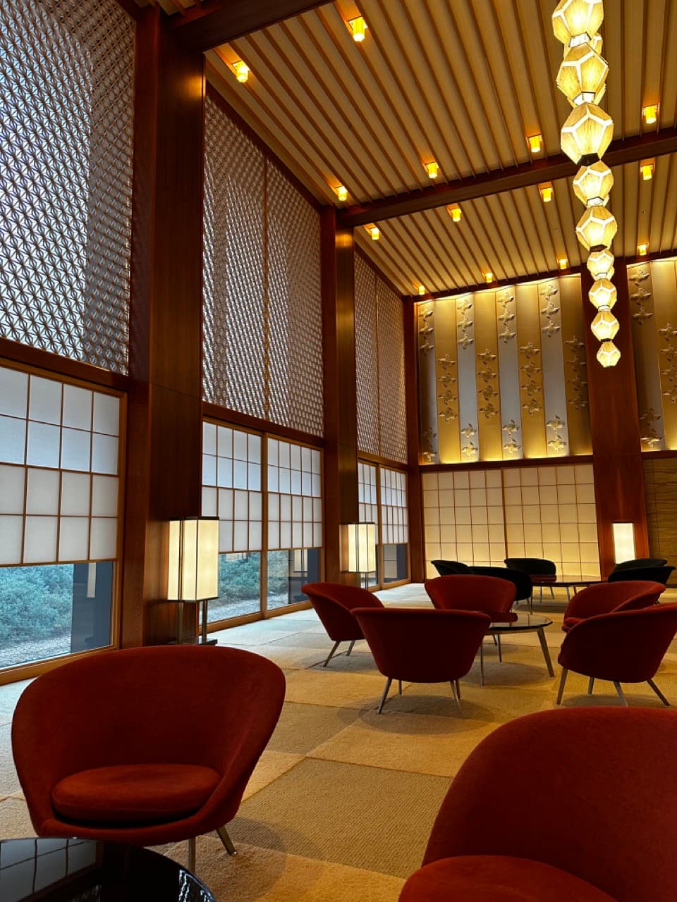 A view of the Okura Tokyo hotel lobby with beige carpet, maroon lounge chairs, overhead lamps provide calm lighting and white blinds allow natural light to flow in.