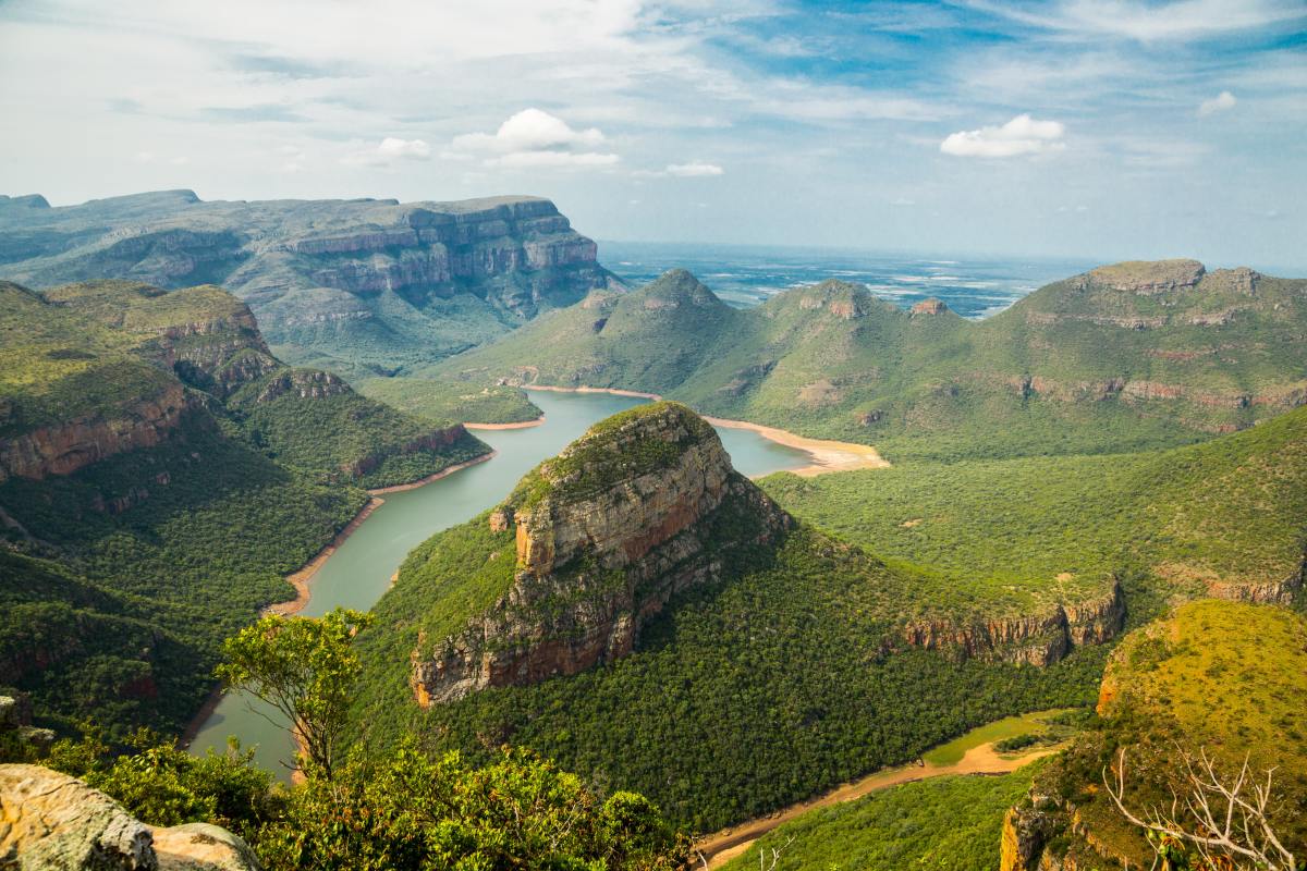 Mountains and rock formations surrounded by greenery and vegetation next to large river in Blyde River Canyon, South Africa.