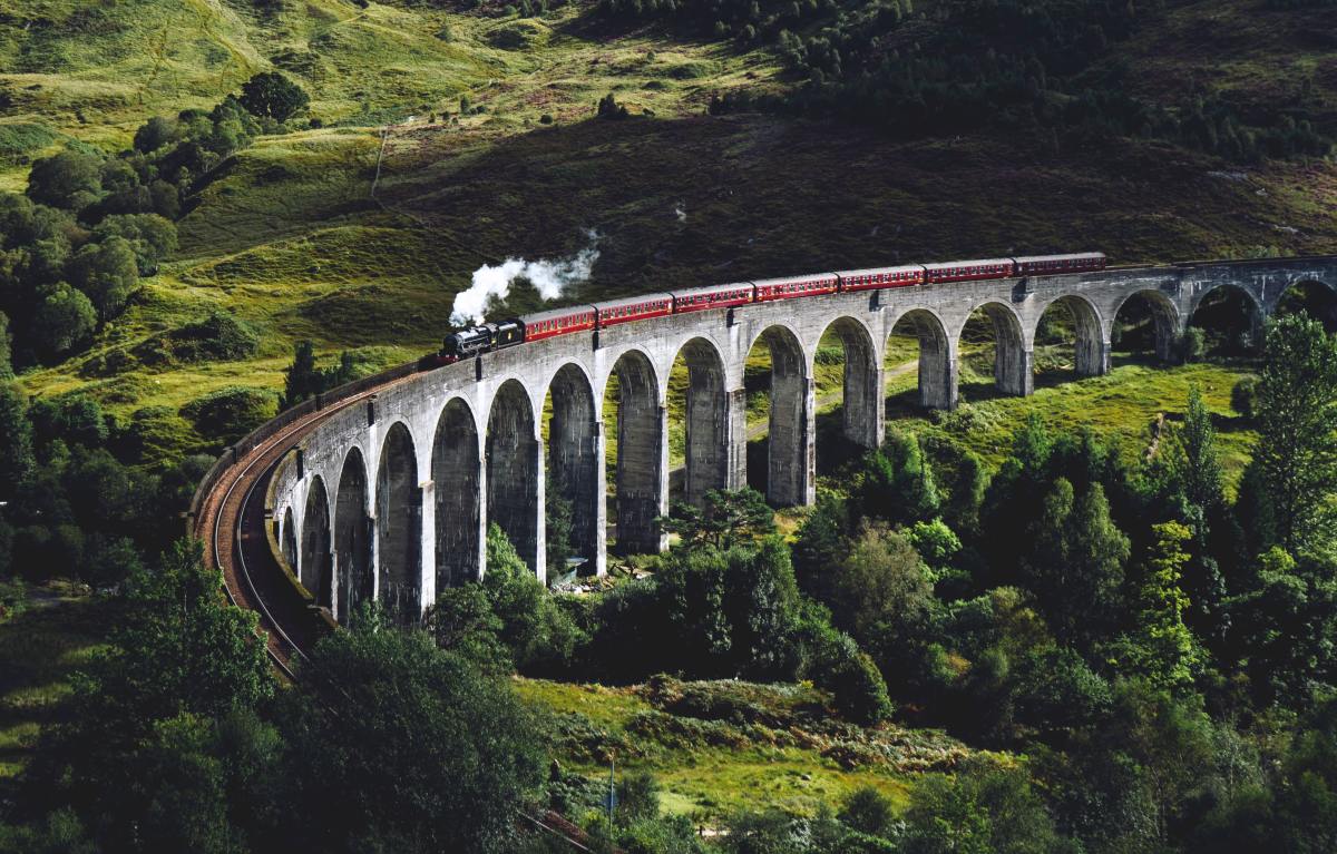 Train travels on bridge surrounded by lush green hills dotted with trees in Scotland