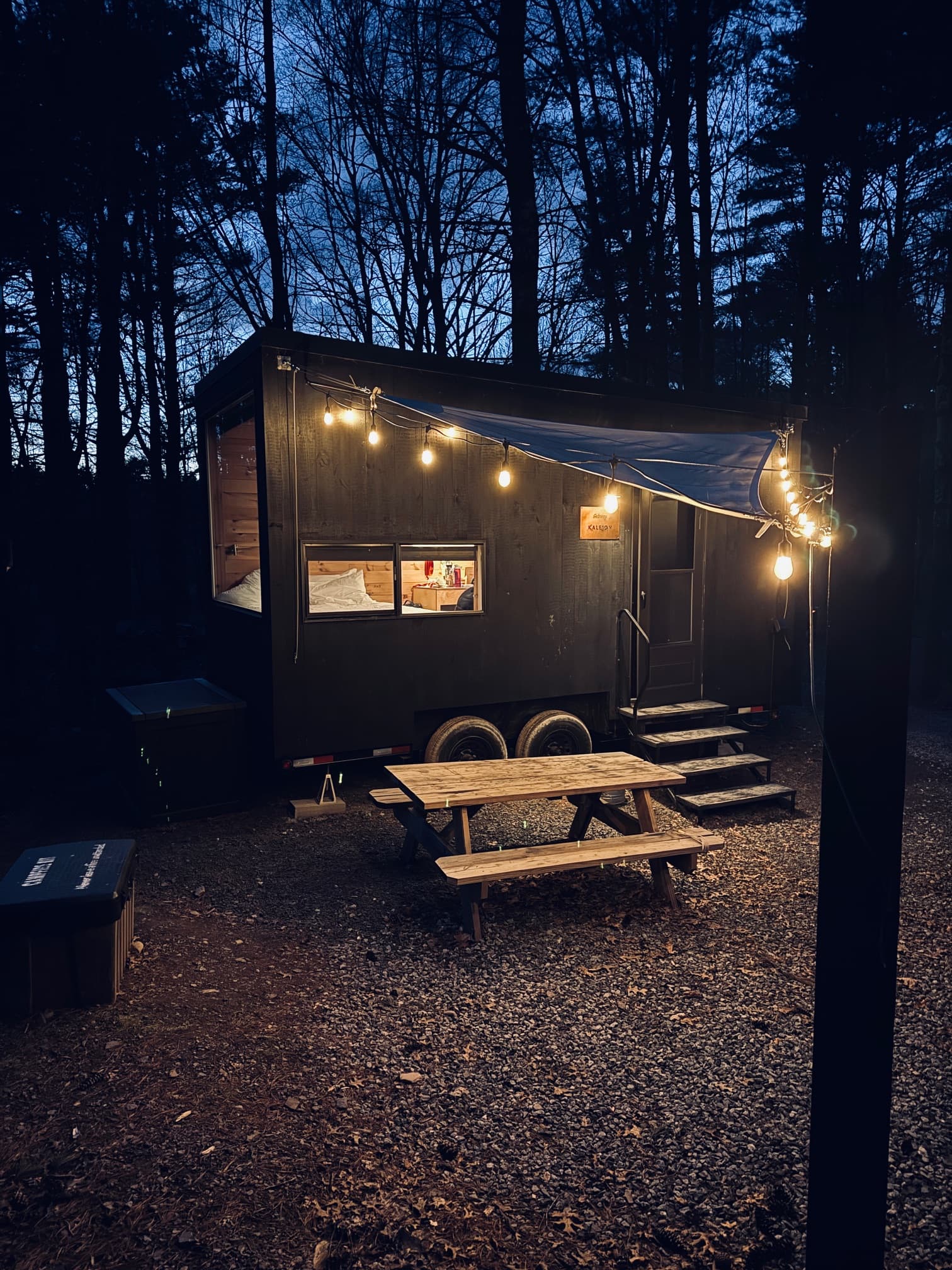 A trailor in the woods with a string of lights illuminating it, and a picnic table in front at night.