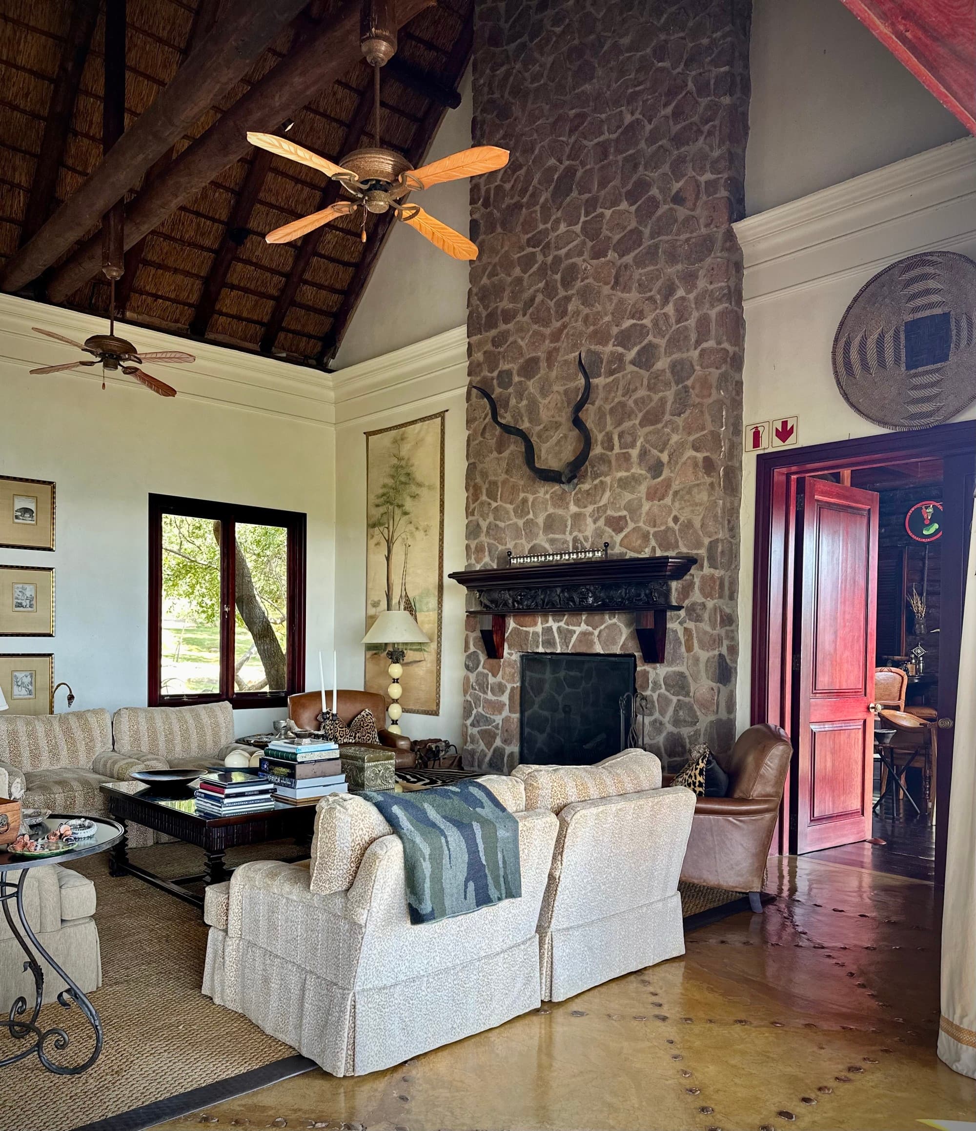 An interior view of a room with a large stone fireplace, mounted antlers, a ceiling fan, striped couches, tables, lamps, art, tiled flooring and red doors beneath a wood beamed ceiling.