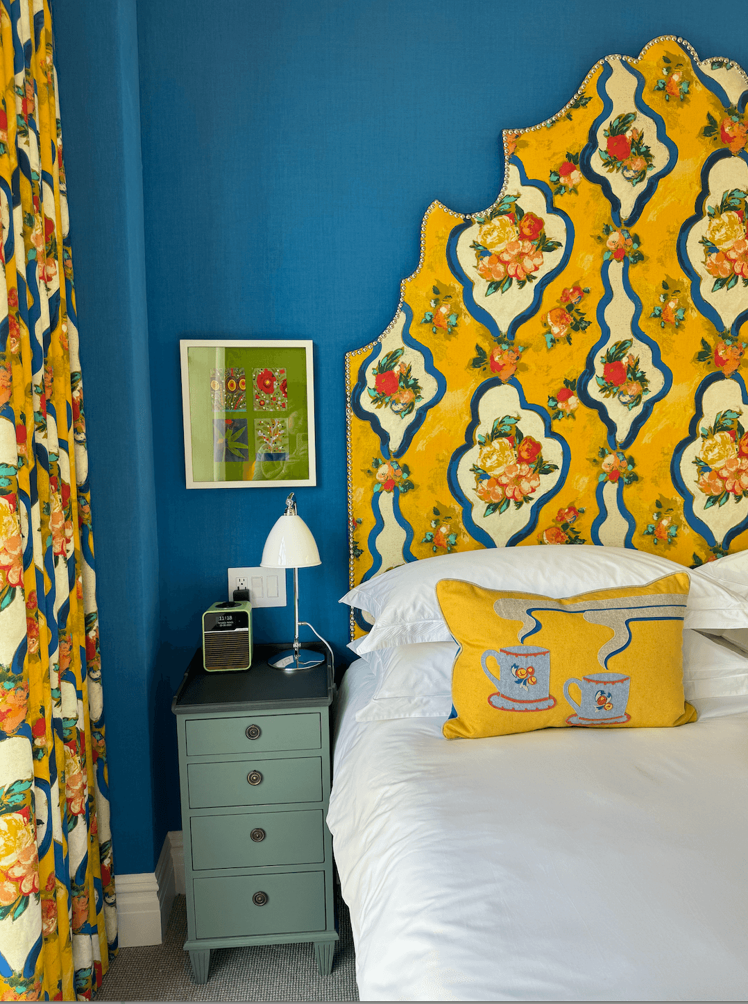A hotel suite with a colorfully patterned headboard, white bedding, blue wall and teal nightstand with a lamp sitting on top of it and piece of artwork hanging behind it.